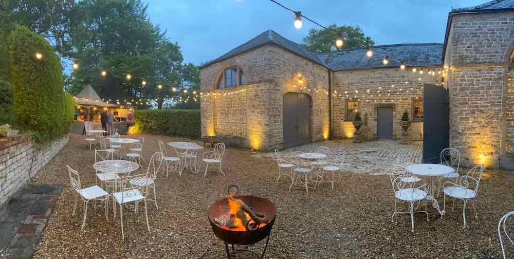 Dusk Elegance at Pennard House Coach House Wedding Venue: The exterior, adorned with festoon lighting, embraces the enchanting twilight. Gravel beneath adds rustic charm, while the gentle glow of a fire-pit imparts warmth. A festoon lighting canopy zigzags across the space, creating a magical ambiance for unforgettable celebrations.
