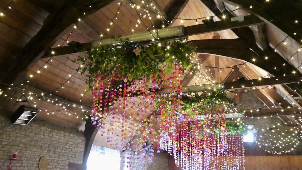 Mells Barn Enchantment: Inside the charming Mells Barn, the ceiling is adorned with a magical display of fairy lights, casting a warm glow. Delicate decorative petals dangle gracefully from above, creating an ethereal atmosphere that adds a touch of enchantment to the rustic interior