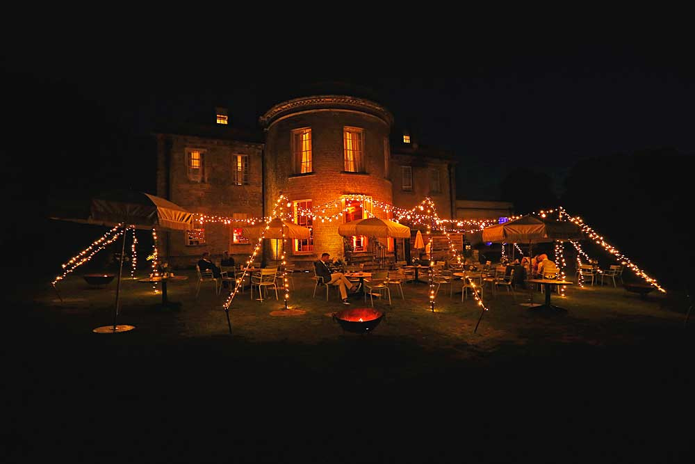 Nighttime Tranquility: On the Babington House Bar Terrace, a man sits beneath a captivating lighting canopy adorned with festoon and fairy lights. The exterior scene glows with a warm and enchanting ambiance, providing a serene setting for relaxation under the night sky.