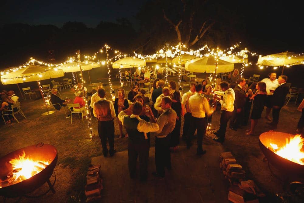 Nighttime Revelry: A wedding lighting canopy creates a mesmerizing scene, accentuated by large fire-pits. Wedding guests mingle under the vast night sky, basking in the warm glow of lights. The atmosphere is one of celebration, where the magic of the night adds to the enchantment of the occasion.