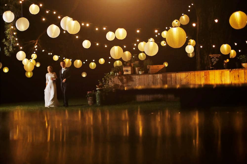 Dramatic Tree Illumination: The scene unfolds with a dramatic display of festoon lighting and glowing globes adorning a majestic tree. To the left, a bride and groom stroll together, creating a magical and enchanting ambiance under the captivating lights.