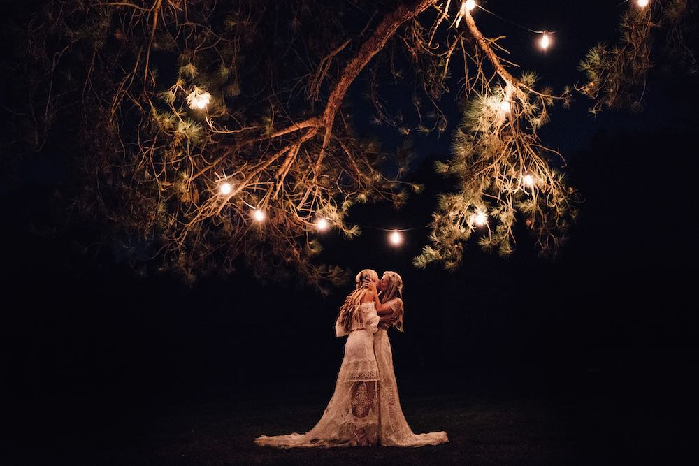 Magical Union: Under the enchanting glow of Edison vintage lights on a serene night, two brides share a tender kiss beneath a magical tree, creating a timeless and romantic moment that echoes the charm of the evening.