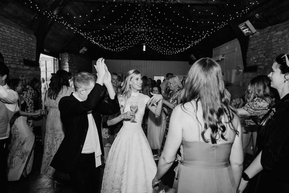Timeless Elegance: In the black and white capture of a stone barn interior, the bride stands at the heart of the frame, surrounded by friends. Overhead, a delicate fairy-light canopy adds a touch of enchantment, creating a timeless and magical ambiance within the rustic setting.