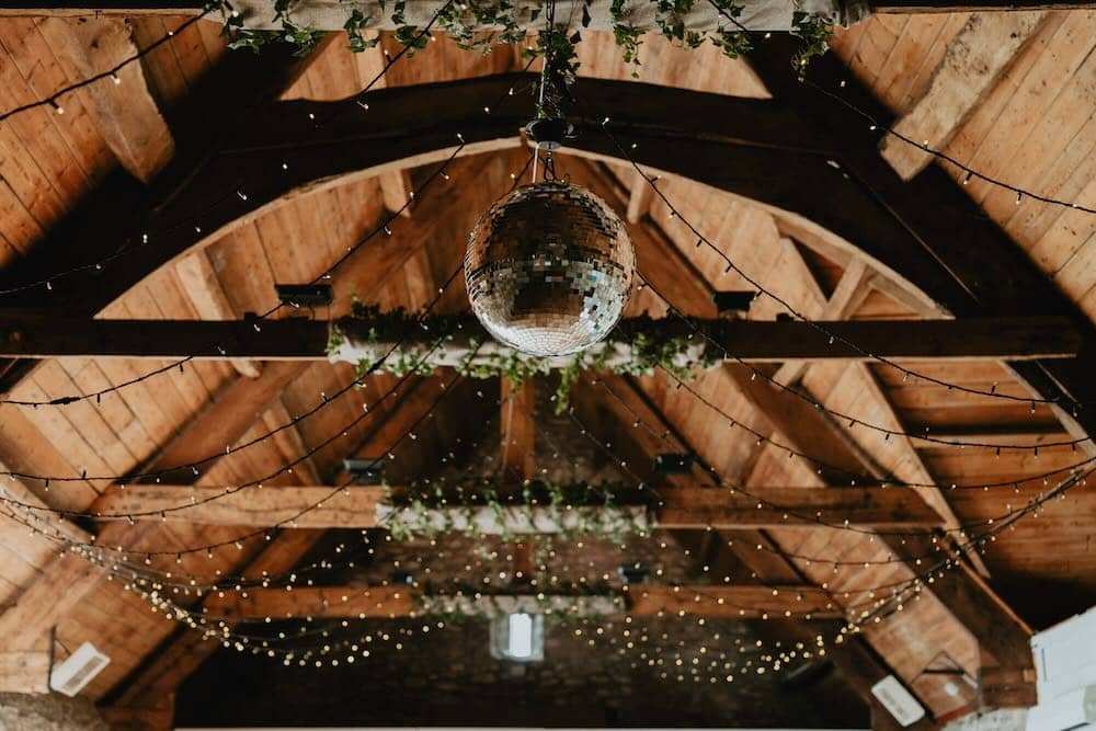 Rustic Elegance: Inside the barn, wooden beams and a charming ceiling set the scene. A mesmerizing mirrorball takes center stage, surrounded by the whimsical glow of fairy lights. Ivy gracefully wraps around the lights on the beams, creating a warm and enchanting ambience in this rustic and elegant space.