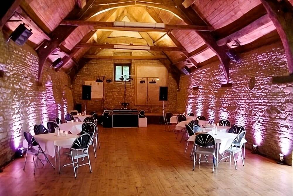 Mells Barn Radiance: Within the rustic charm of Mells Barn in Somerset, pink LED up-lights cast vibrant hues, painting the stone walls with playful shades. Tables and chairs line each side, bathed in the gentle glow. At the end, a DJ setup awaits, creating a lively and colorful atmosphere within this captivating venue.