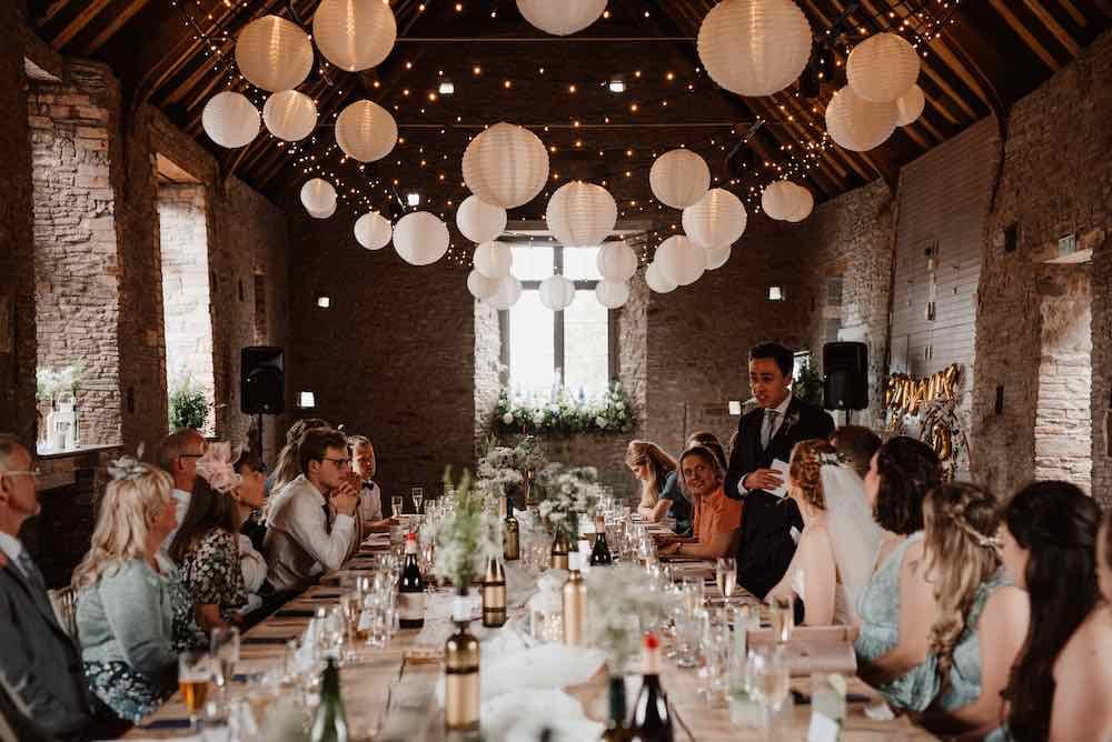 Daytime Elegance in the Stone Barn: Inside the rustic stone barn, a wedding reception unfolds in the soft daylight. The groom stands at the forefront, delivering a heartfelt speech, while guests are seated at a long table, immersed in the joyous atmosphere of this special celebration.