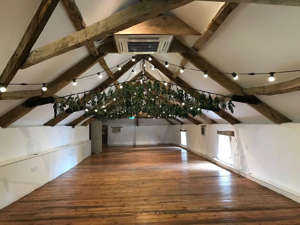 Pennard House Wedding Ambiance: In the empty coach house at Pennard House, Somerset, wedding lighting takes center stage. Festoon lights adorned with foliage drape elegantly, infusing the space with a magical ambiance that sets the tone for a romantic celebration.