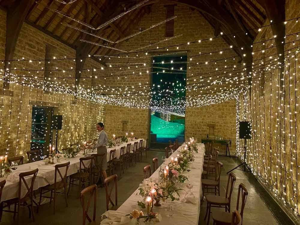 Rustic Stone Barn Setting: Two elongated tables adorned with chairs, embellished with flowers and candles. Overhead, a magical tunnel of fairy lights gracefully traverses the length of the barn, casting a warm and enchanting glow.