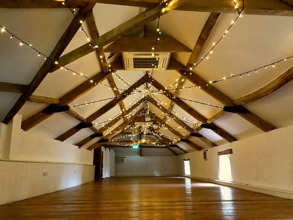 Attic Barn Tranquility: Inside the white-walled attic barn, wooden beams add rustic charm to the space. The large, empty wooden floor awaits the magic of celebration. Above, a mesmerizing display of fairy lights gracefully covers the ceiling, creating a warm and tranquil ambiance in this uniquely styled interior.