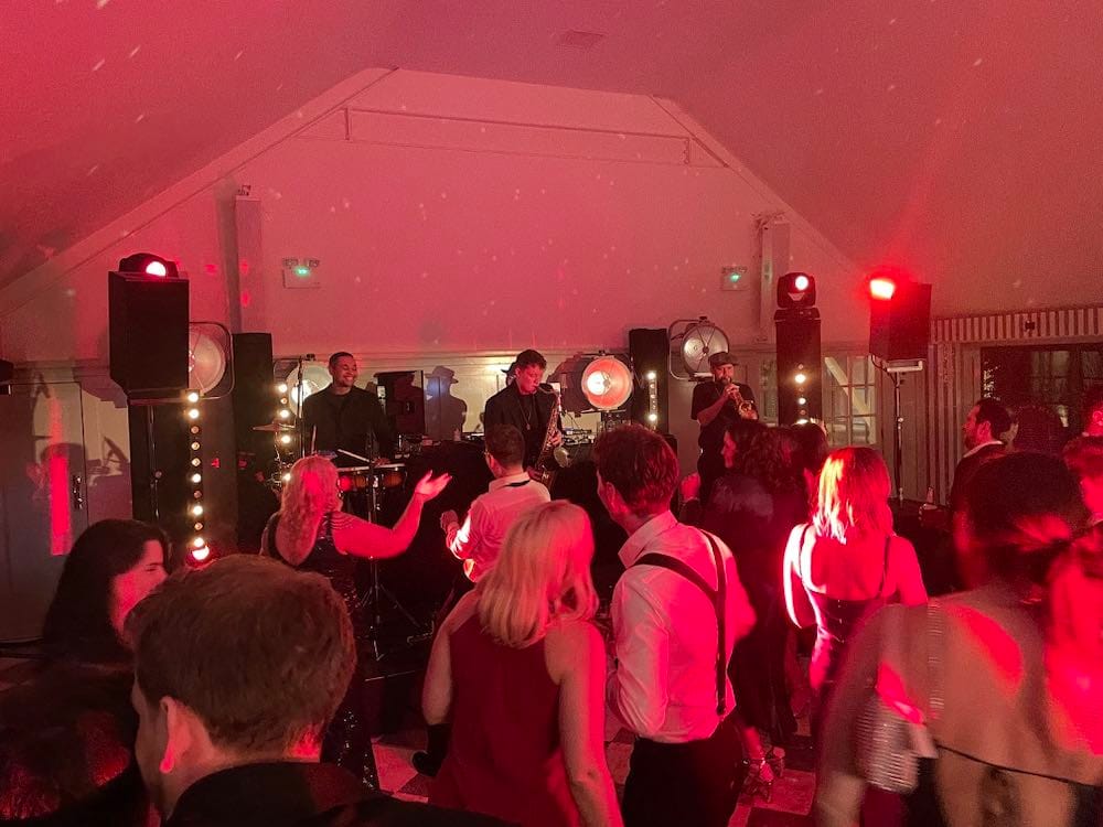Stage lighting transforms Kin House as a live band takes the spotlight, bathed in a captivating red glow. The audience, appreciative and immersed, enjoys the performance amidst the dynamic interplay of lights, creating a vibrant and memorable musical experience.