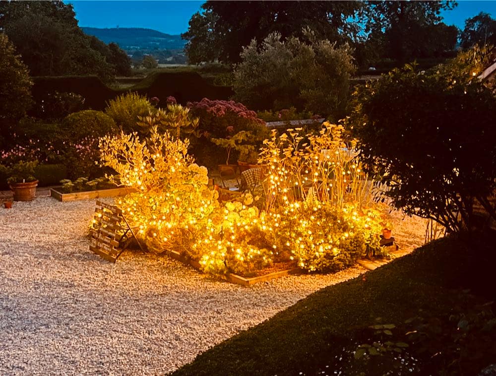 Evening Charm in the English Garden: As dusk settles, the exterior reveals an enchanting English garden. A vibrant flowerbed takes center stage, adorned with the gentle glow of fairy lights, casting a magical ambiance over the gravel-covered ground. The scene captures the serene beauty of twilight in the garden.