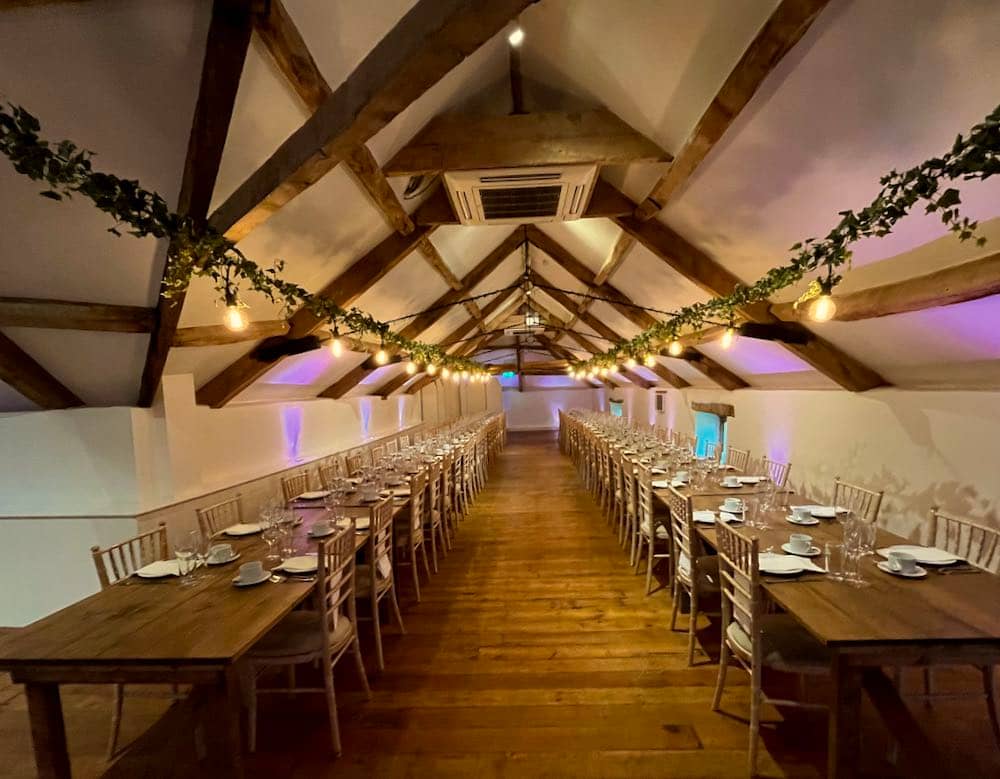 Rustic Chic: Inside the barn, white-painted walls complement the warmth of wooden beams. Two long tables stretch down the length of the room, adorned with Edison Vintage bulbs running along the center of each, casting a soft and nostalgic glow that enhances the rustic charm of the setting.