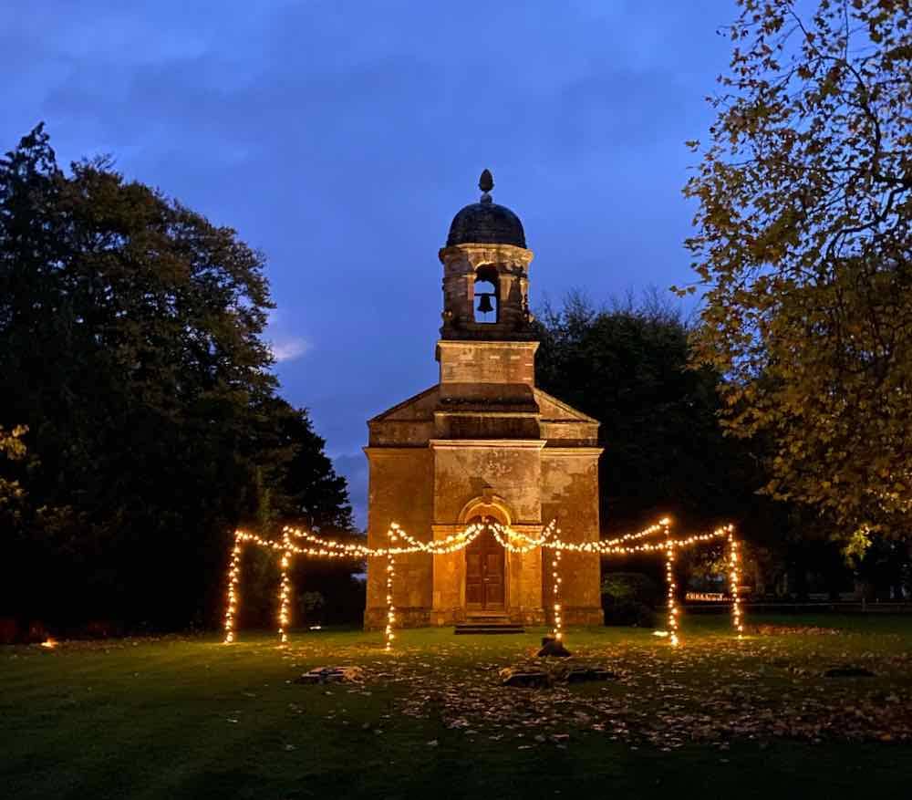 Dusk's Graceful Canopy: As night gently descends, a stone church takes center stage. Trees stand sentinel on either side, and from the church door, a fairy-light canopy gracefully fans out, suspended on upright poles. The scene captures the enchantment of dusk, creating a magical ambiance around the timeless church setting.