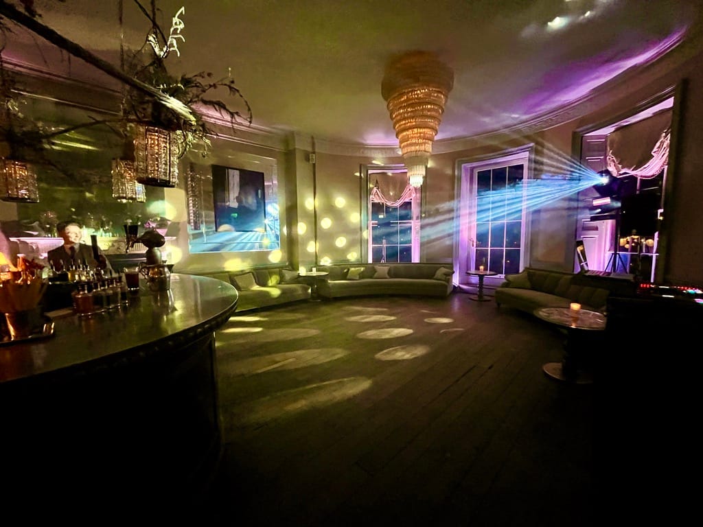 Nighttime Elegance: In the bar's evening ambiance, three large windows illuminate the wooden floor, softly bathed in pink lighting. Dance-floor lighting on the walls adds a touch of sophistication, setting the stage for a stylish gathering as the anticipation builds for the arrival of wedding guests.